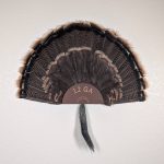 Kabash Outdoors American steel turkey fan display hanger. Fan holder is rustic brown and looks like the end of a 12ga shotgun shell. It is holding a beautiful turkey tail an beard.